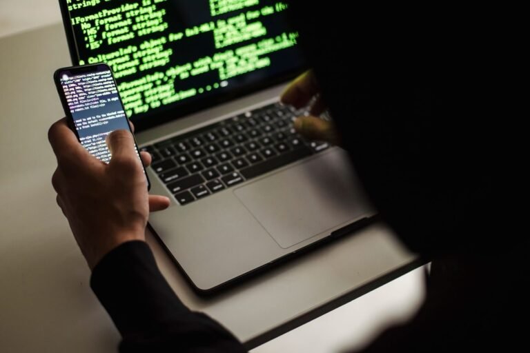 Persona con un teléfono móvil en la mano frente a un computador portátil con código en la pantalla, en un ambiente oscuro